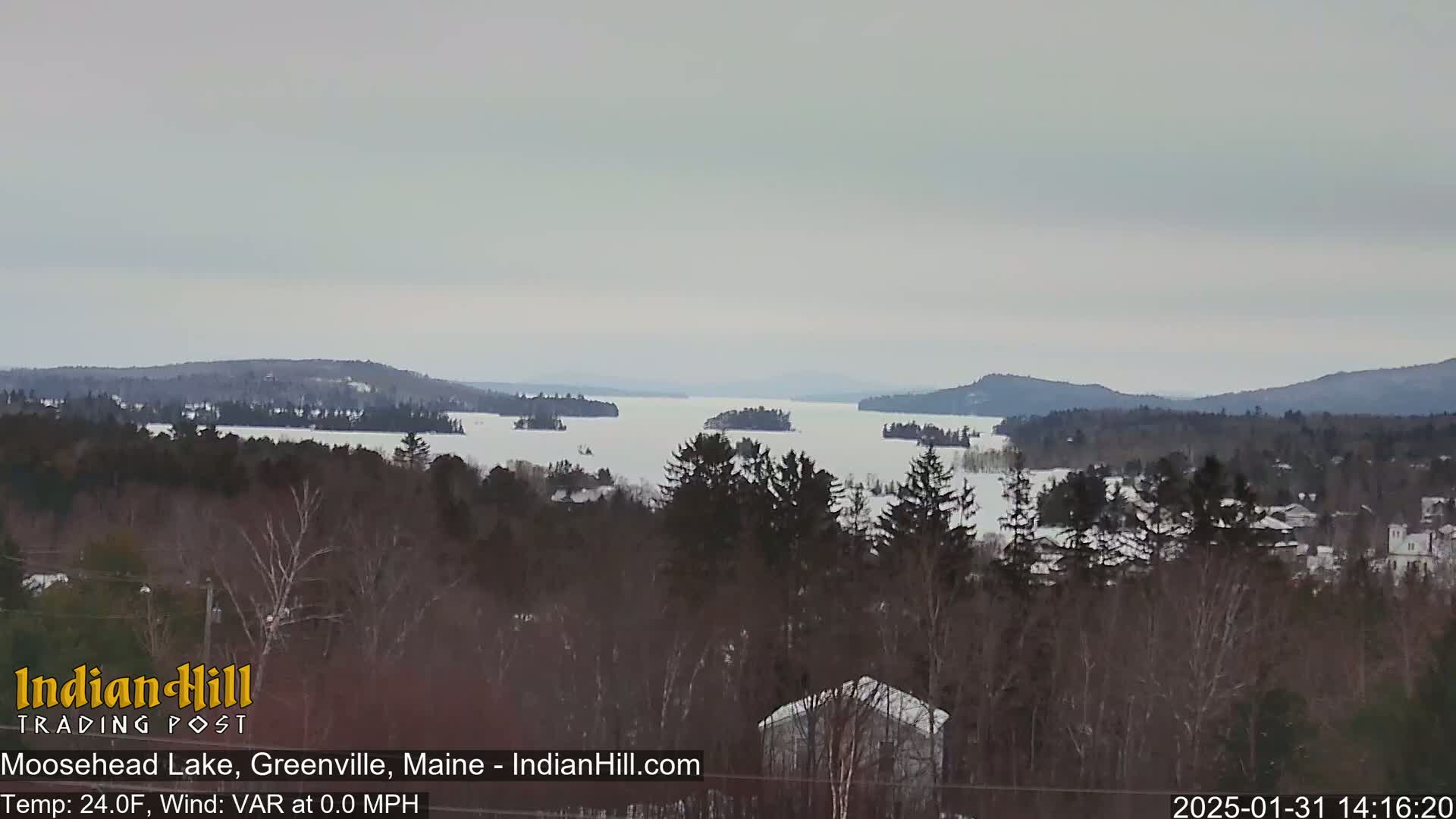 A snow-covered, frozen lake is seen from a distance, surrounded by a snow-covered forest and distant hills under an overcast sky.