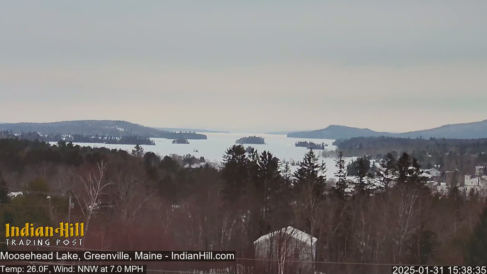 A snow-covered lake with several islands is visible from a wooded, snow-dusted hilltop under an overcast sky.