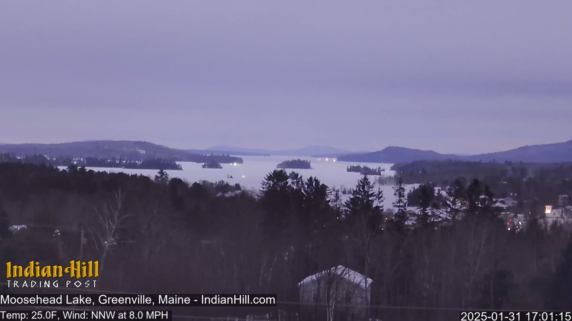 A twilight view shows a partially frozen lake surrounded by snow-covered trees and distant mountains under a dusky sky.