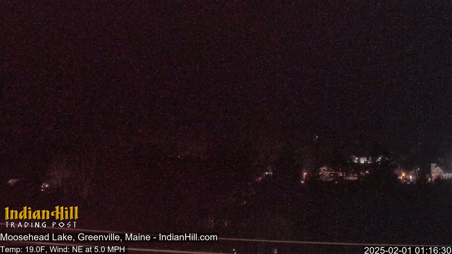 A nighttime long shot of sparsely lit buildings and trees under a dark sky, with a northeast wind at 5.0 mph and a temperature of 19 degrees Fahrenheit.