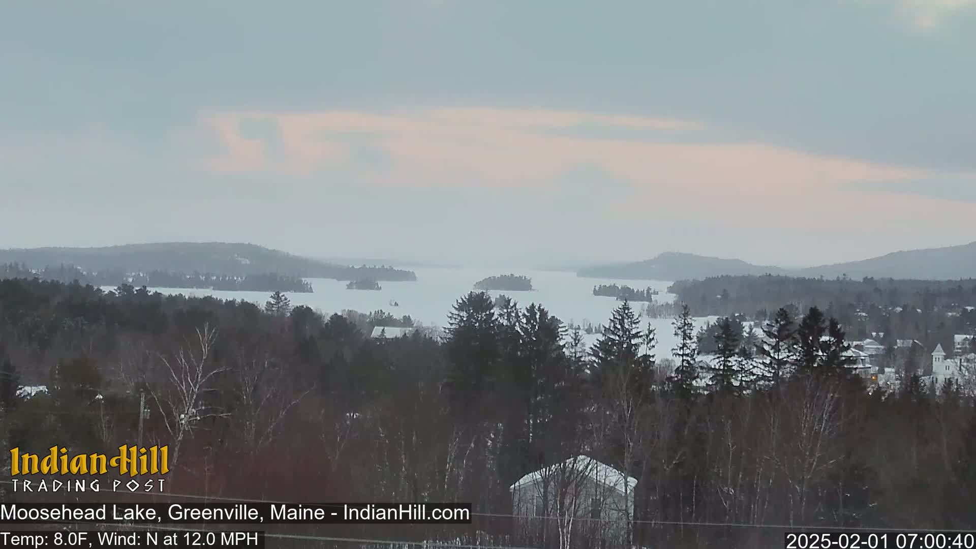 A snow-covered lake with several small islands is visible from a vantage point overlooking a snow-covered forest under an overcast sky.