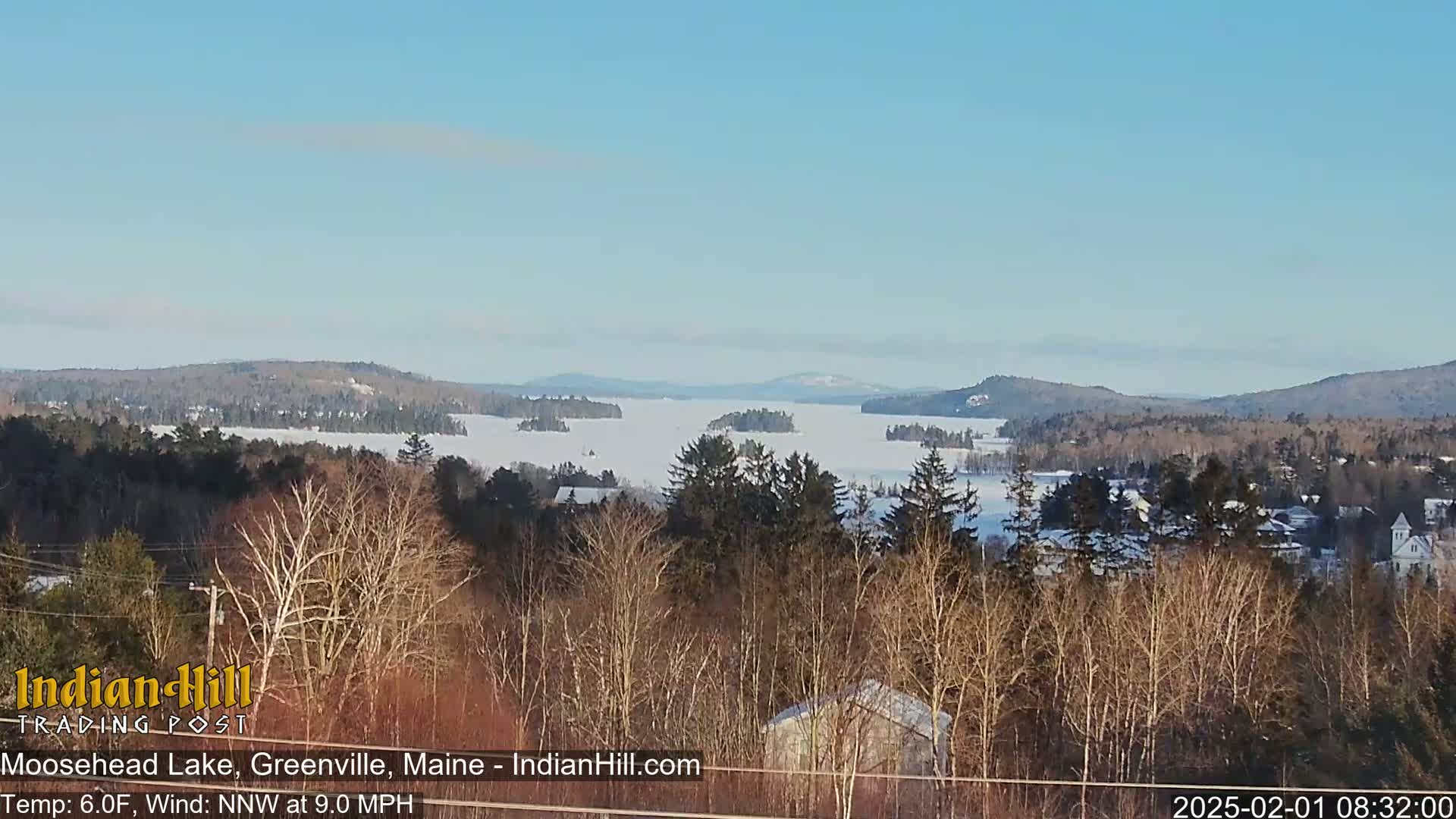 A snow-covered lake stretches to distant, snow-dusted mountains under a clear, bright sky, viewed from a vantage point amongst bare winter trees.
