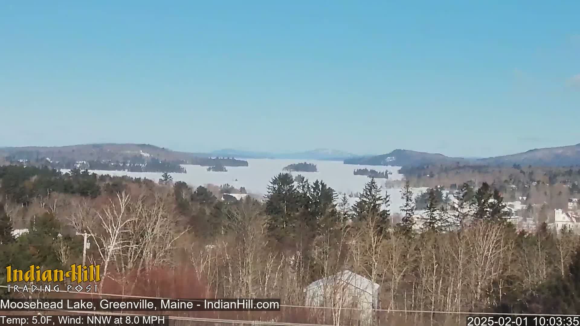 A snow-covered lake stretches into the distance, surrounded by a mix of evergreen and deciduous trees under a clear blue sky.