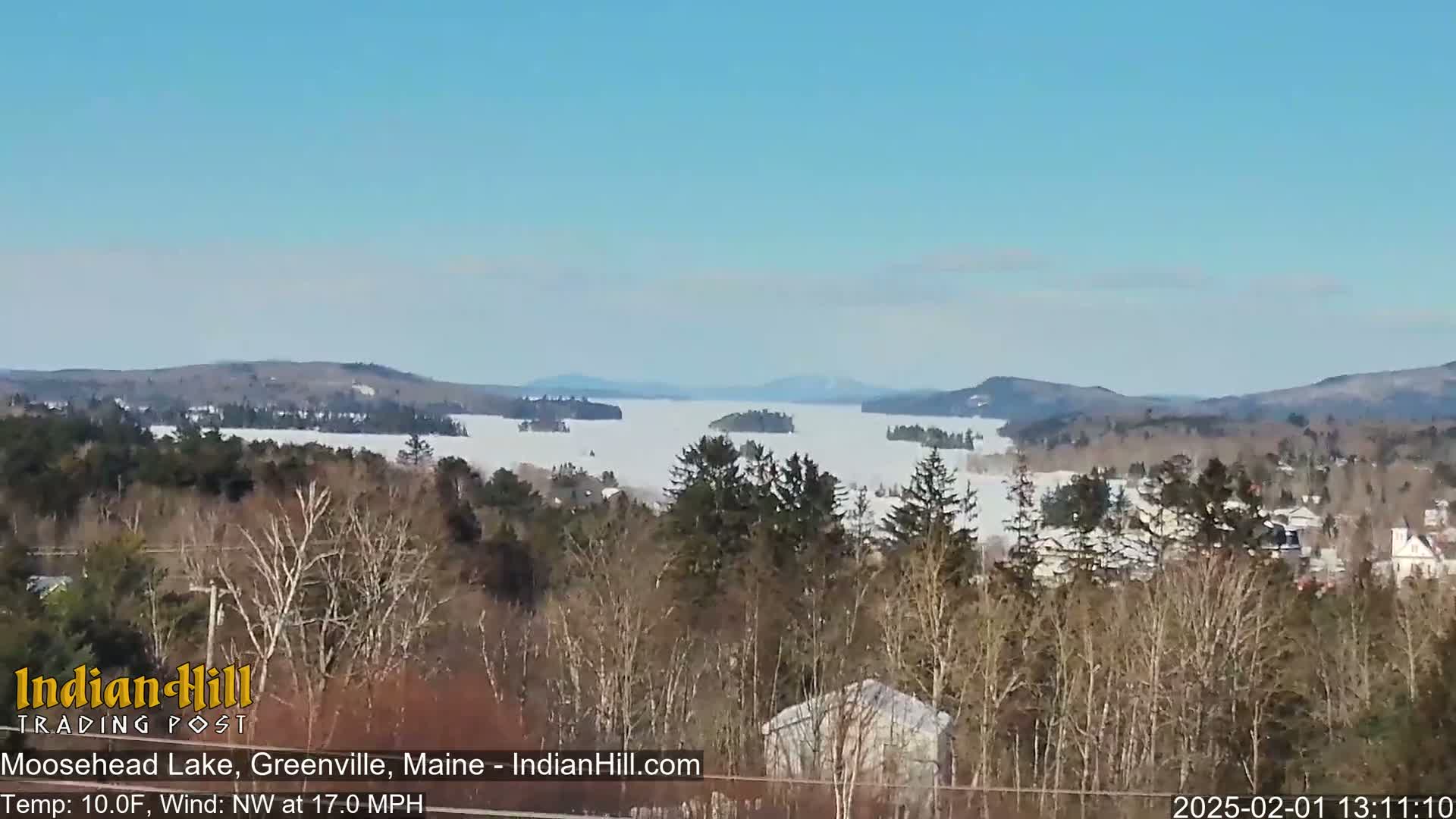 A snow-covered lake with islands and distant mountains is seen from a high vantage point on a clear, cold, and windy day.