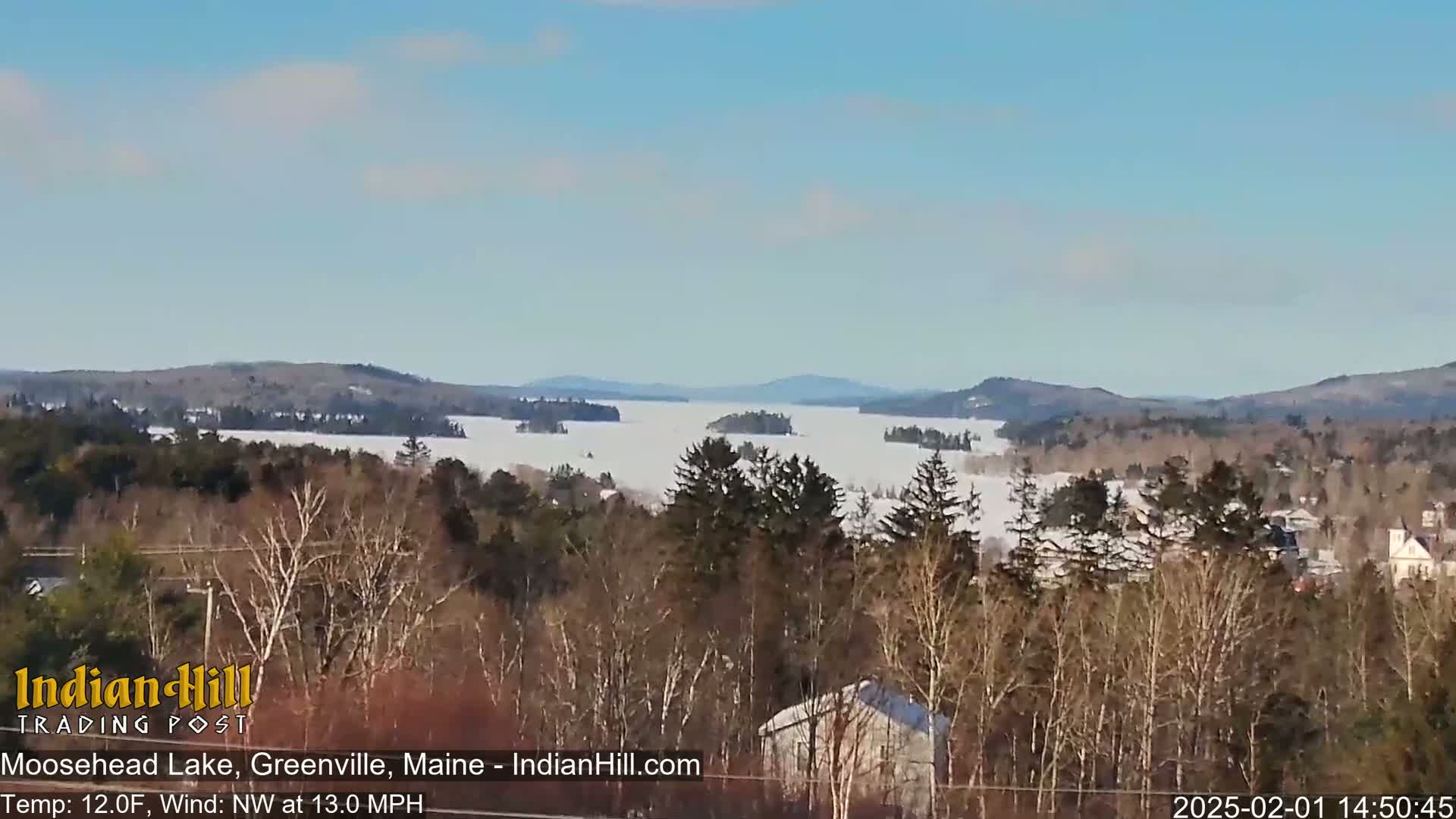 A snow-covered lake stretches between snow-dusted hills under a clear, bright sky, viewed from a wooded hillside.