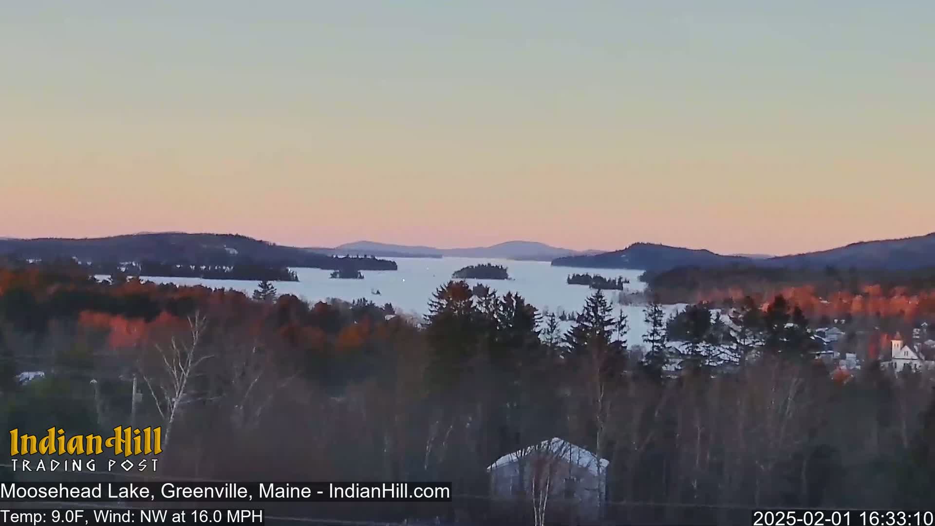 A snow-covered lake is surrounded by dark, forested hills under a pale pink and purple sunset sky.