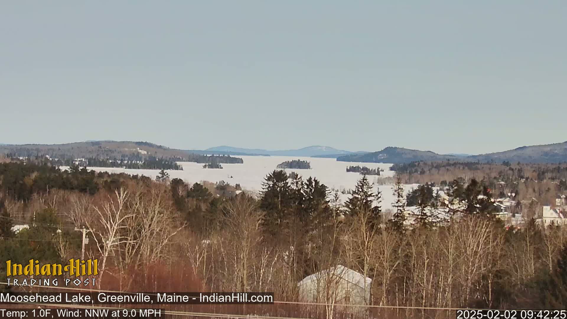 A snow-covered lake with several small islands is visible from a wooded hillside on a cold, windy day.