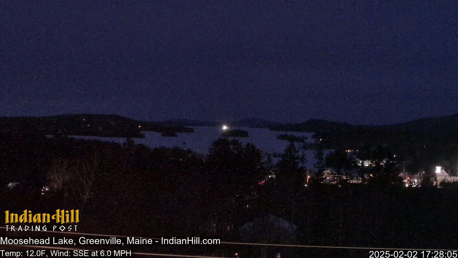 A nighttime view of a snow-covered lake and surrounding wooded areas, under a dark sky with a slight snowfall.