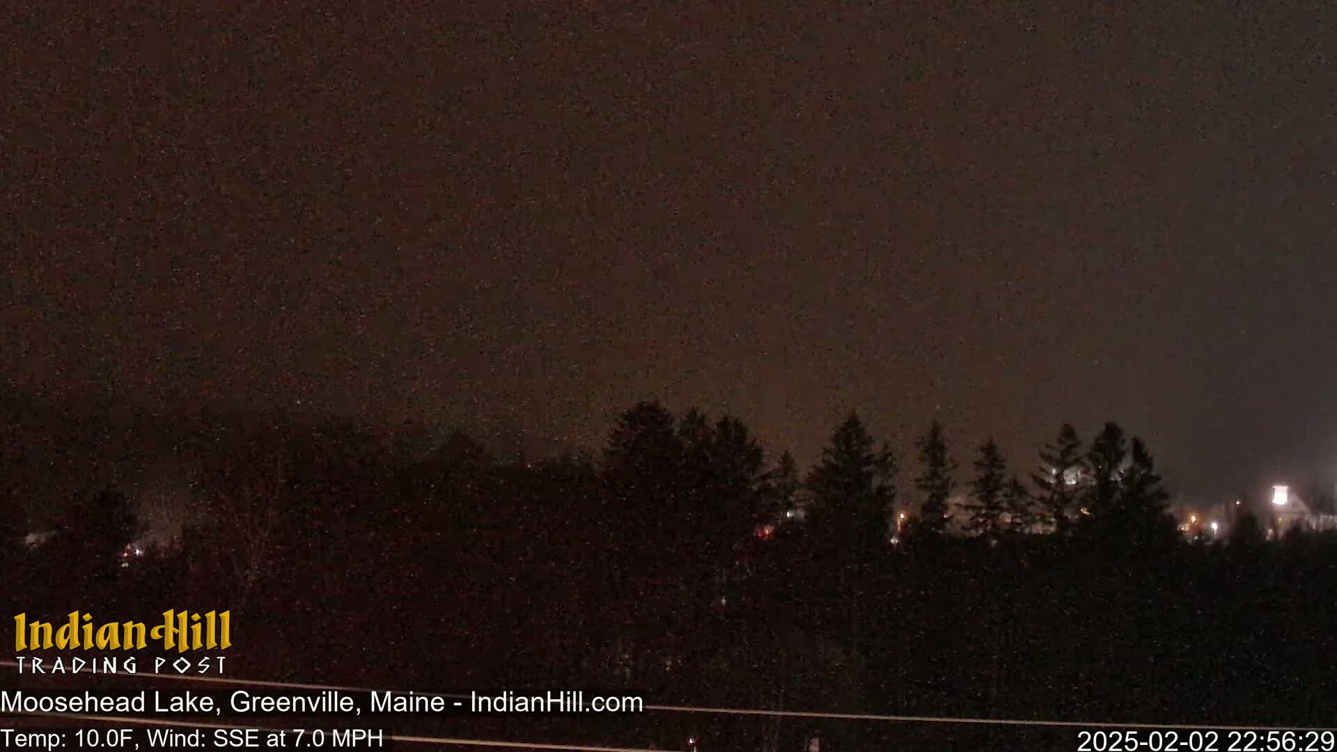 A nighttime view of a snow-covered landscape with a dark sky and a line of evergreens in the distance.