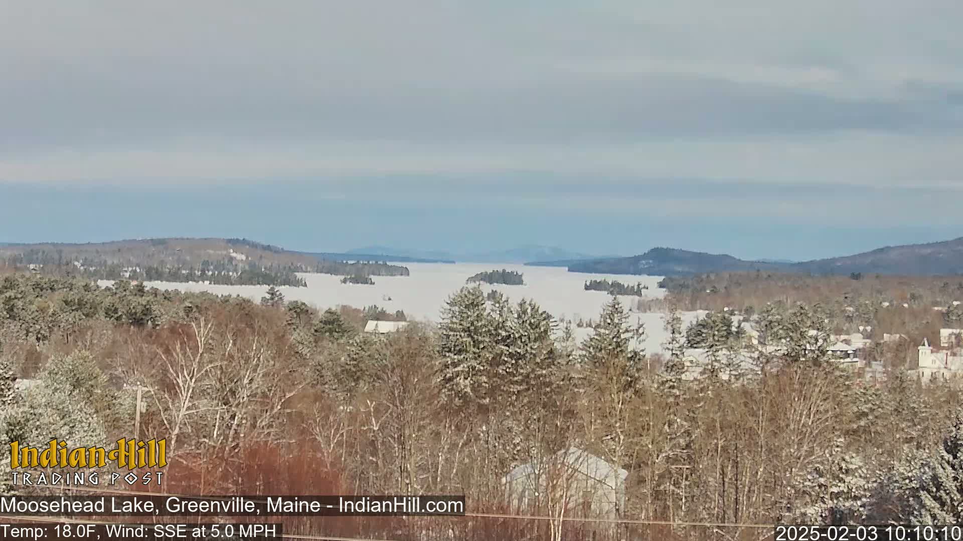 A snow-covered, frozen lake is visible from a snowy, wooded hilltop under a mostly clear sky.