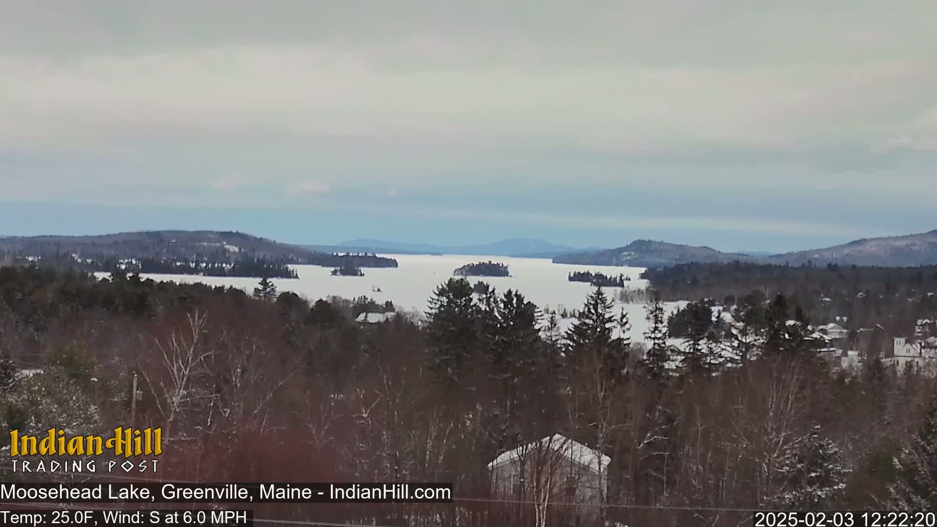 A snow-covered lake with several small islands is visible from a wooded hillside under a cloudy sky.