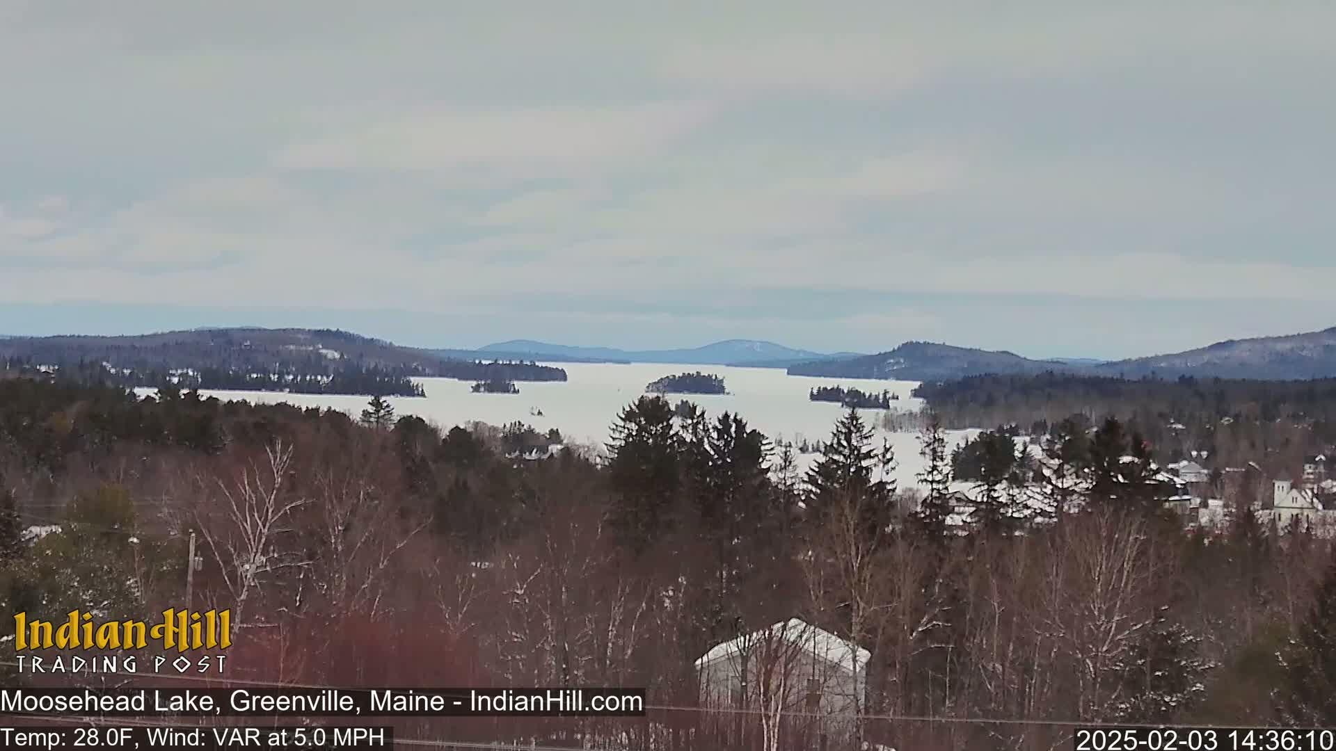 A snow-covered lake stretches to distant, snow-dusted mountains under a cloudy sky.