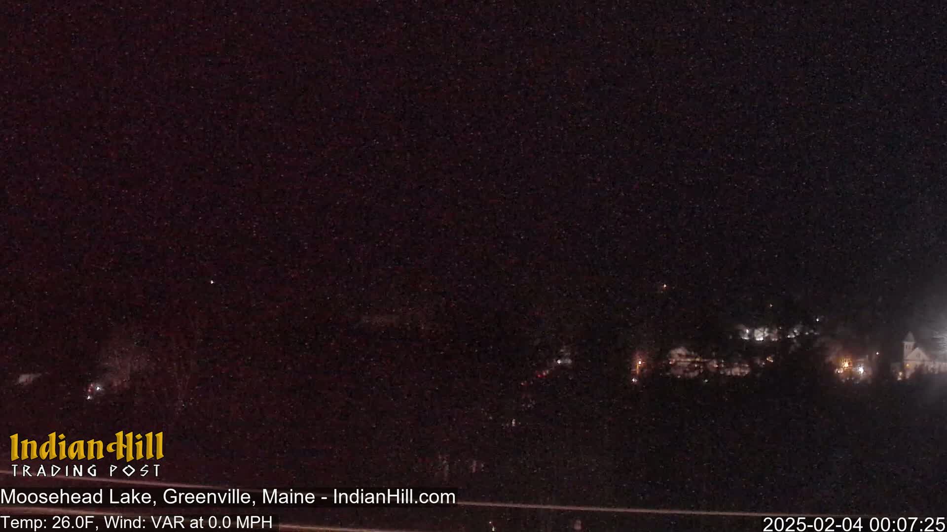 A nighttime, dark view of a sparsely lit lakeside town under a clear, star-filled sky.