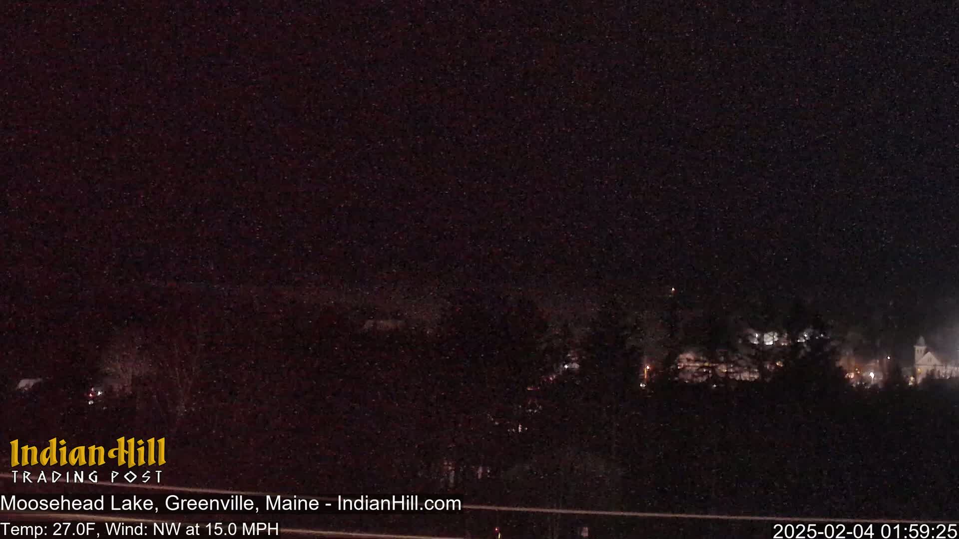 A nighttime, snow-filled scene shows a distant town with faint lights under a dark sky, with a northwest wind blowing at 15 mph and a temperature of 27°F.