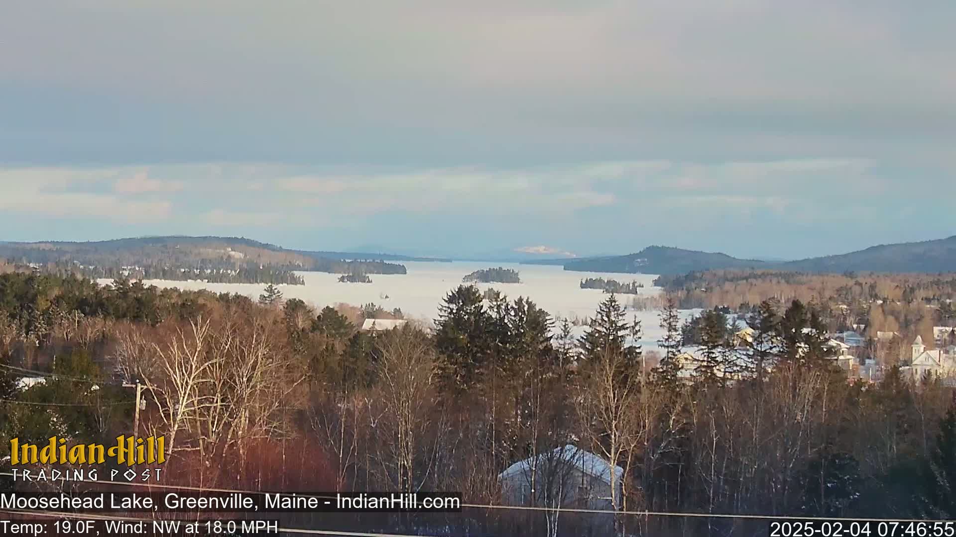 A snow-covered lake stretches to distant, snow-dusted mountains under a mostly cloudy sky, with a foreground of snow-covered evergreen trees and houses.