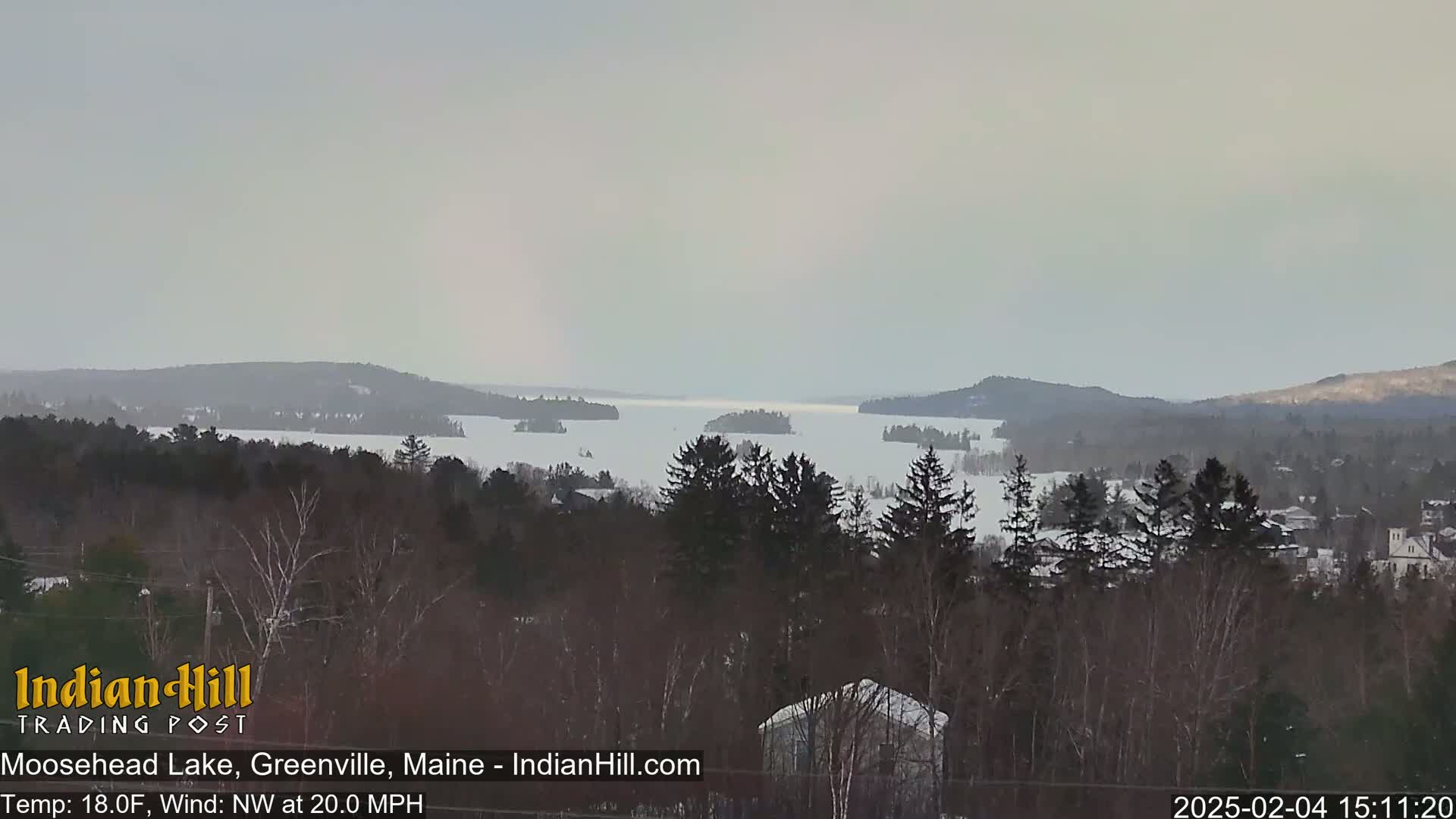 A snow-covered lake with numerous small islands is visible from a wooded hillside under an overcast sky with a brisk northwest wind.