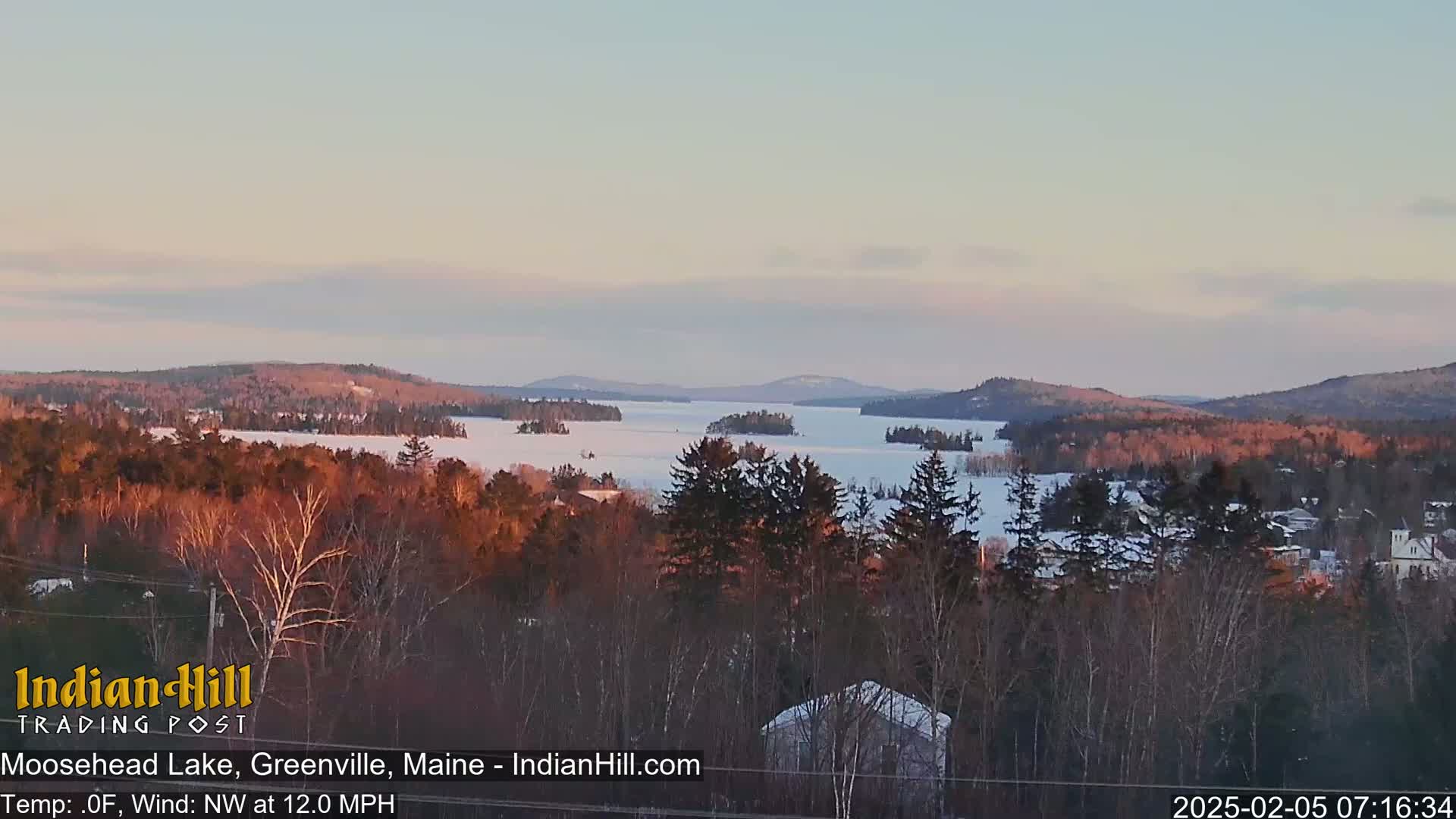 A snow-covered lake stretches to the horizon under a pale pink and blue sky, surrounded by dark, snow-dusted trees and distant hills.