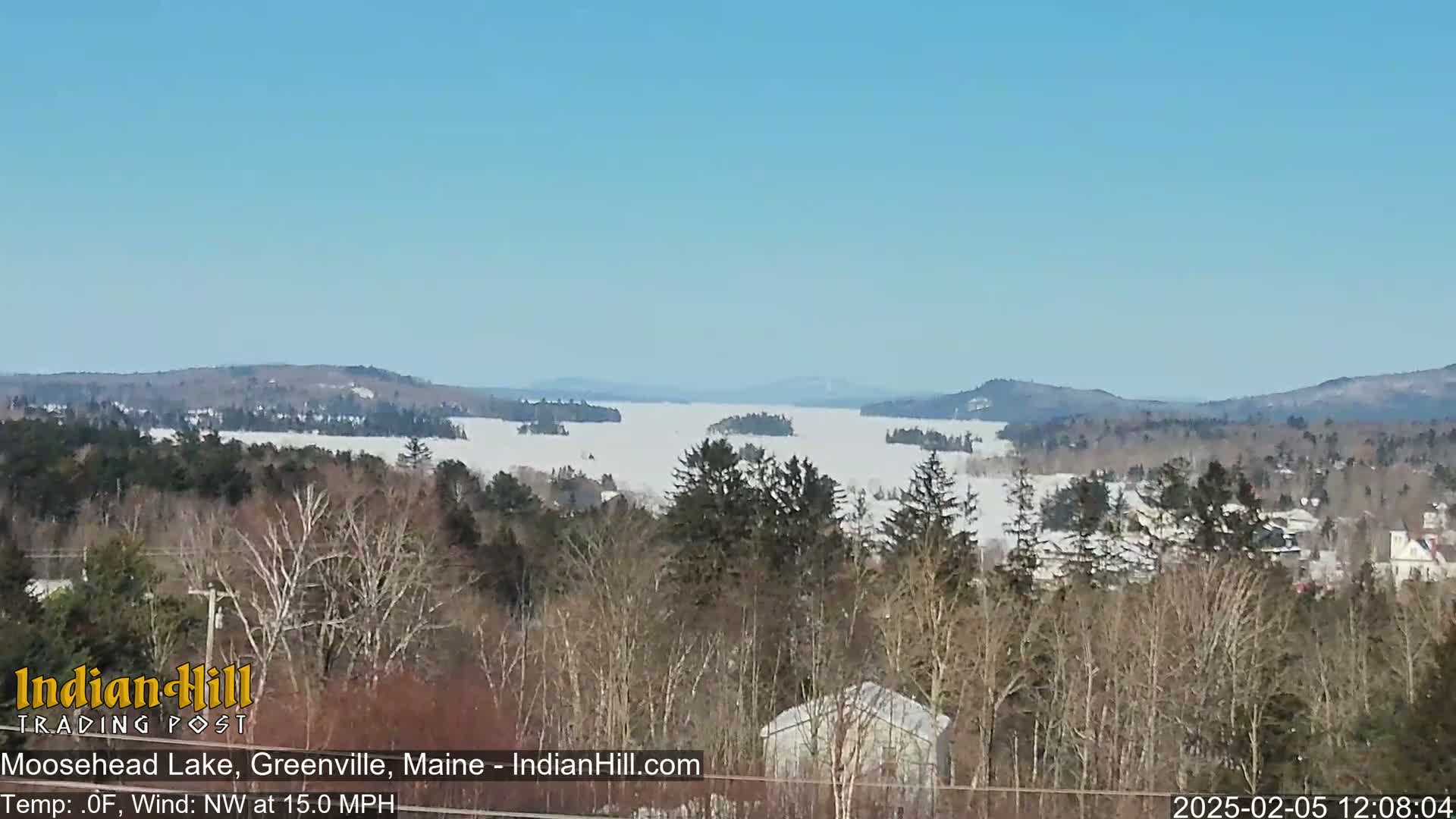A snow-covered lake stretches into the distance under a clear blue sky, framed by leafless trees and distant mountains.