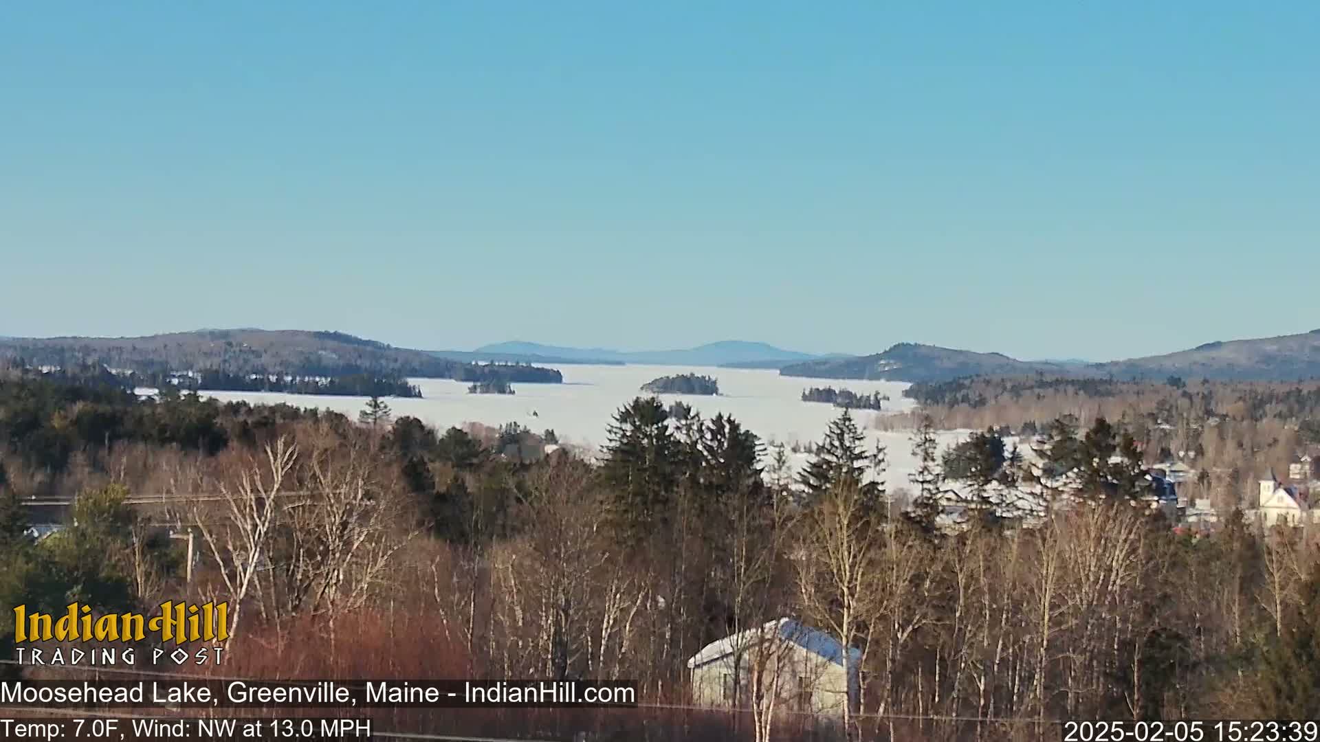 A snow-covered lake stretches out to distant, snow-dusted mountains under a clear blue sky, viewed from a vantage point overlooking a snow-covered forest and a few buildings.