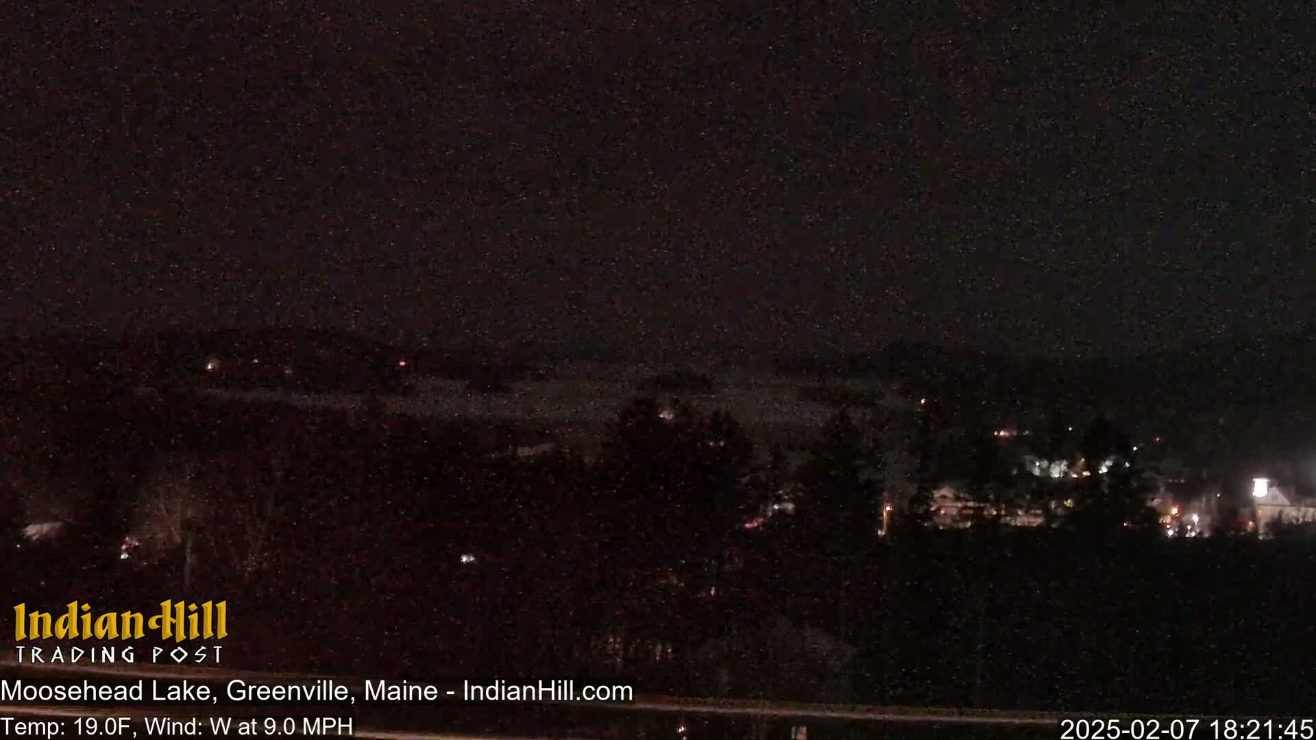 A nighttime view of a lake and town under snowy conditions with a 9.0 mph west wind and a temperature of 19°F.
