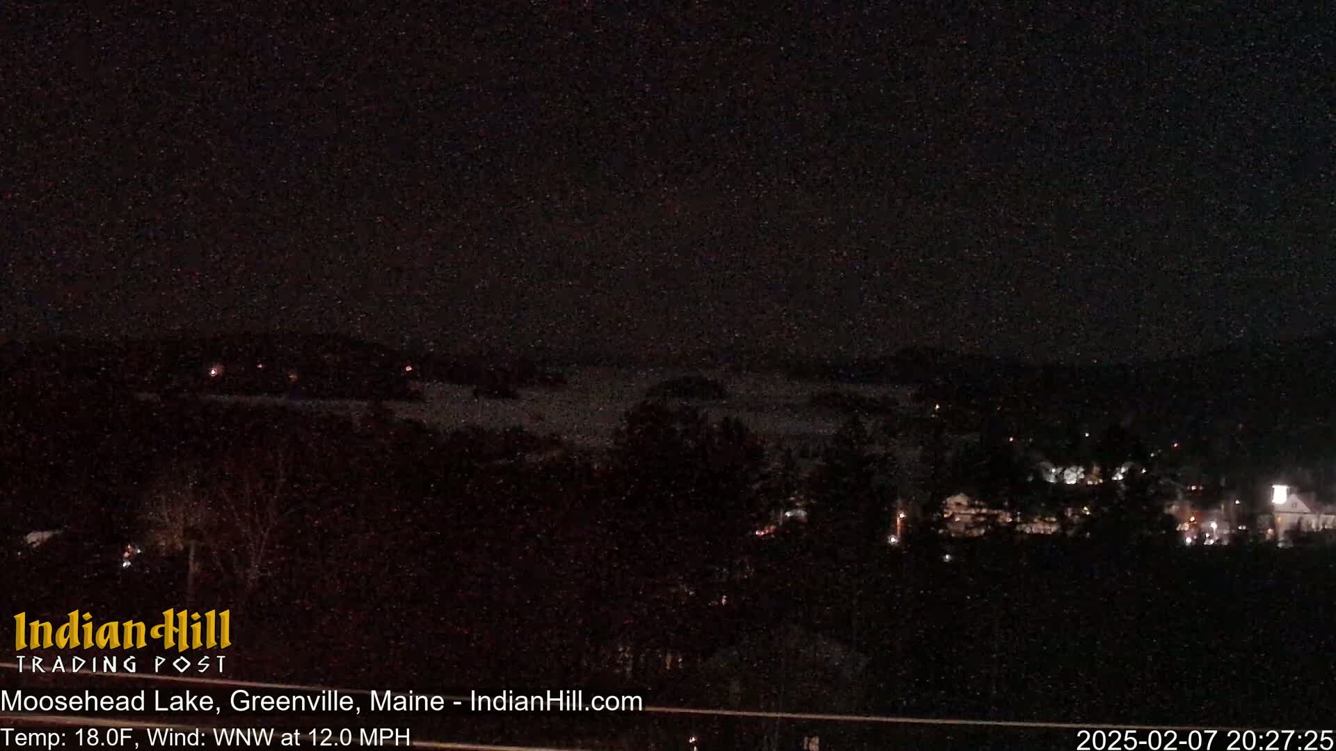 A nighttime view of a lake and sparsely lit shoreline under a dark, clear sky.