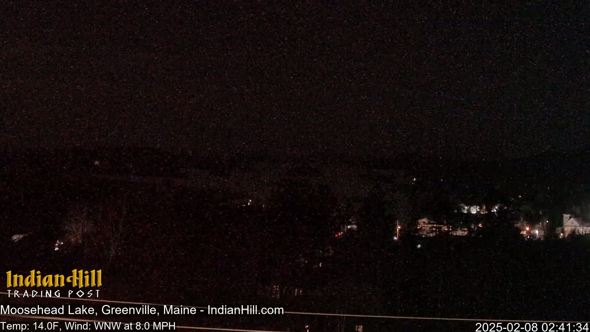 A nighttime long shot of a dark, sparsely lit town under a clear, star-filled sky with a temperature of 14 degrees Fahrenheit and a west-northwest wind of 8 miles per hour.