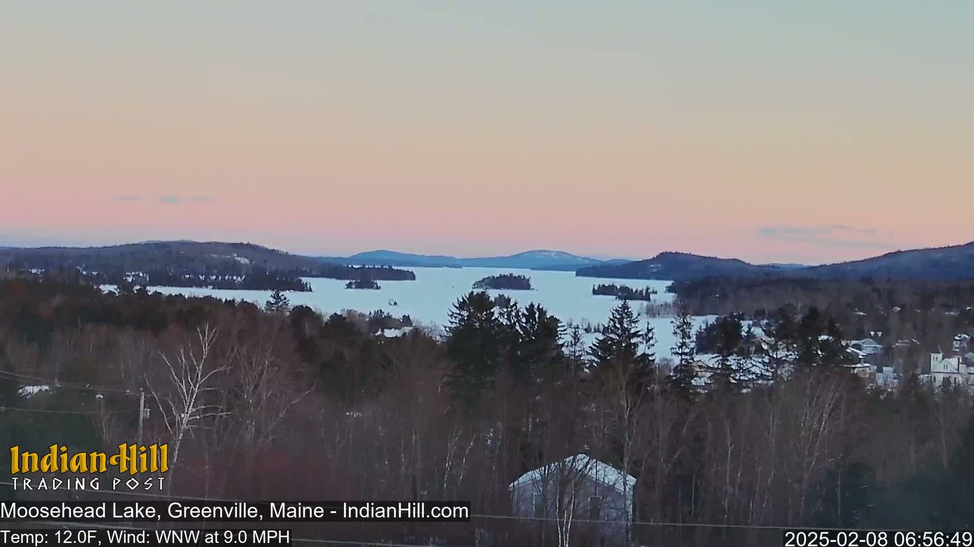A snow-covered lake with islands is visible at sunset, viewed from a wooded hillside under a pale pink sky.