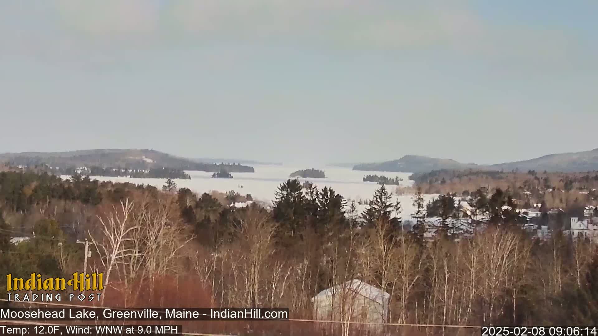 A snow-covered lake stretches to the horizon, framed by a winter landscape of bare trees and distant hills under a light gray sky.