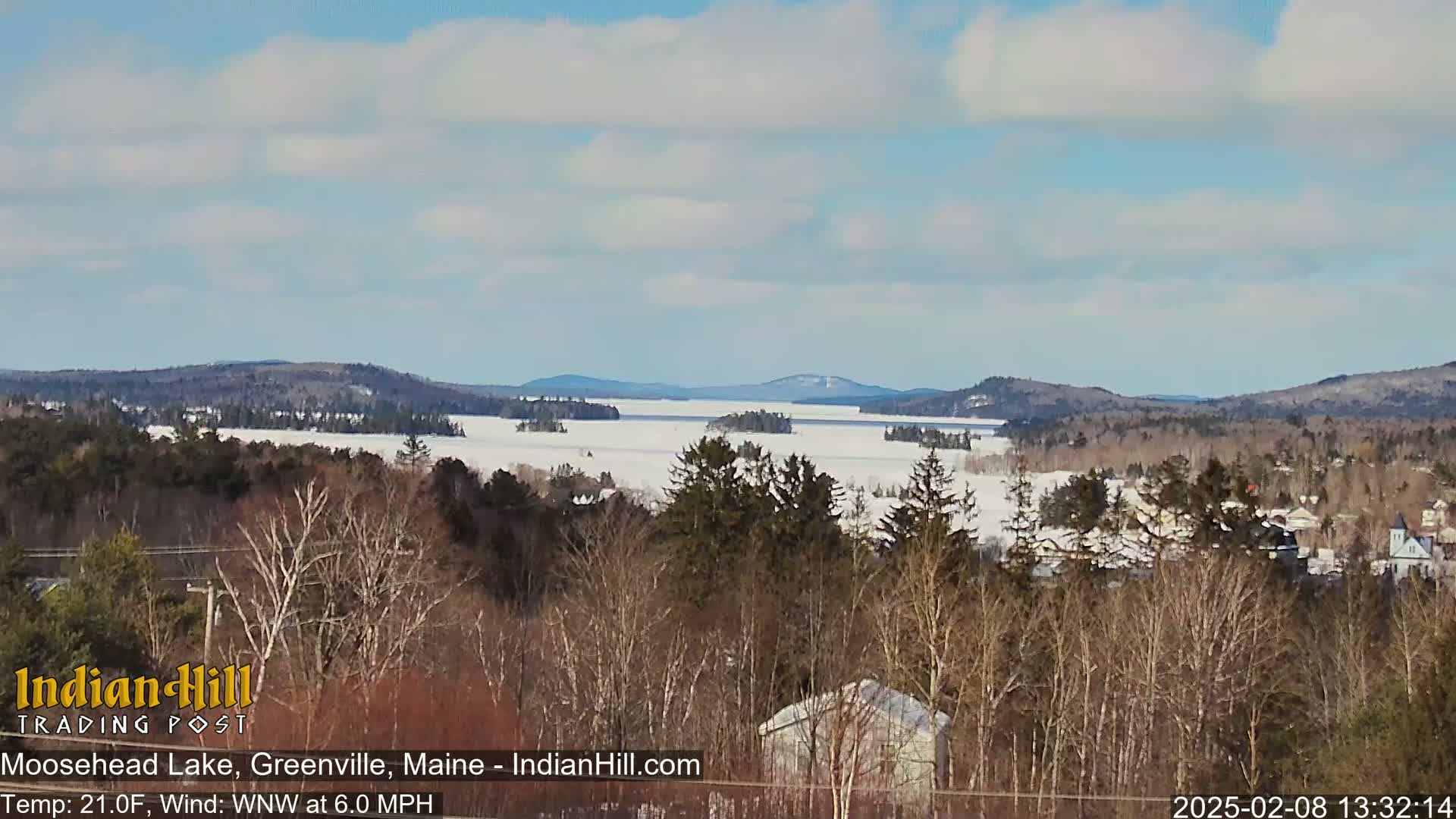 A snow-covered lake stretches to distant, wooded hills under a partly cloudy, bright sky.