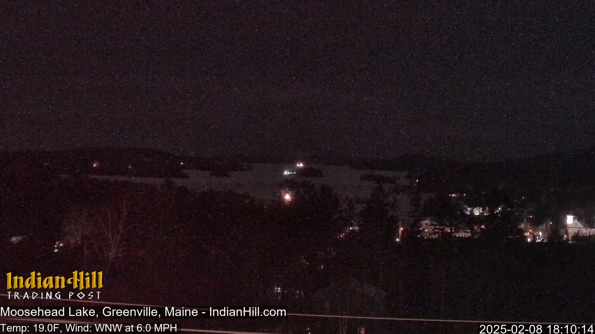A nighttime view of a dark lake with sparsely lit houses and trees along its shores under a clear, dark sky.