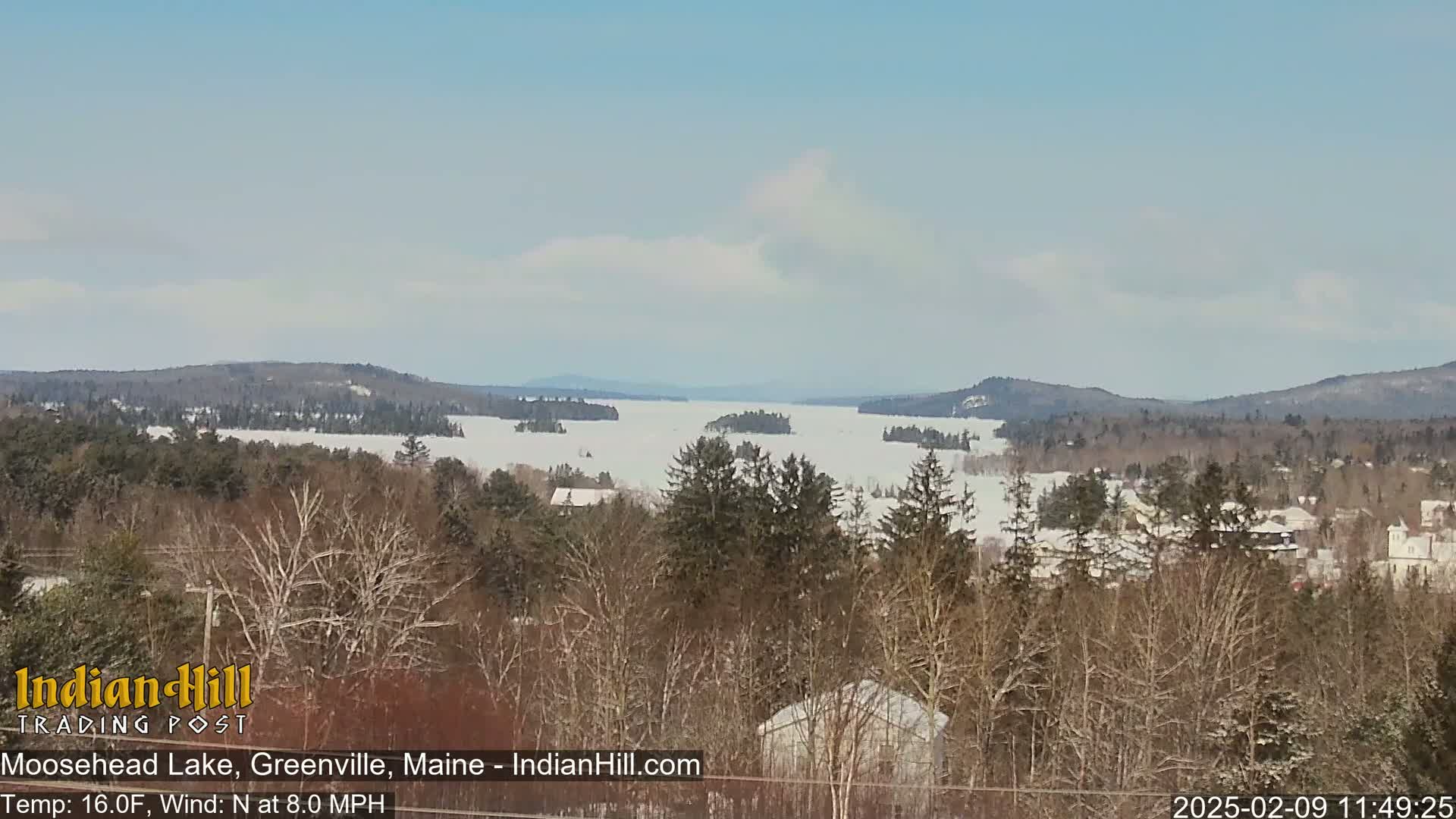 A snow-covered lake stretches to distant, snow-dusted mountains under a partly cloudy sky.