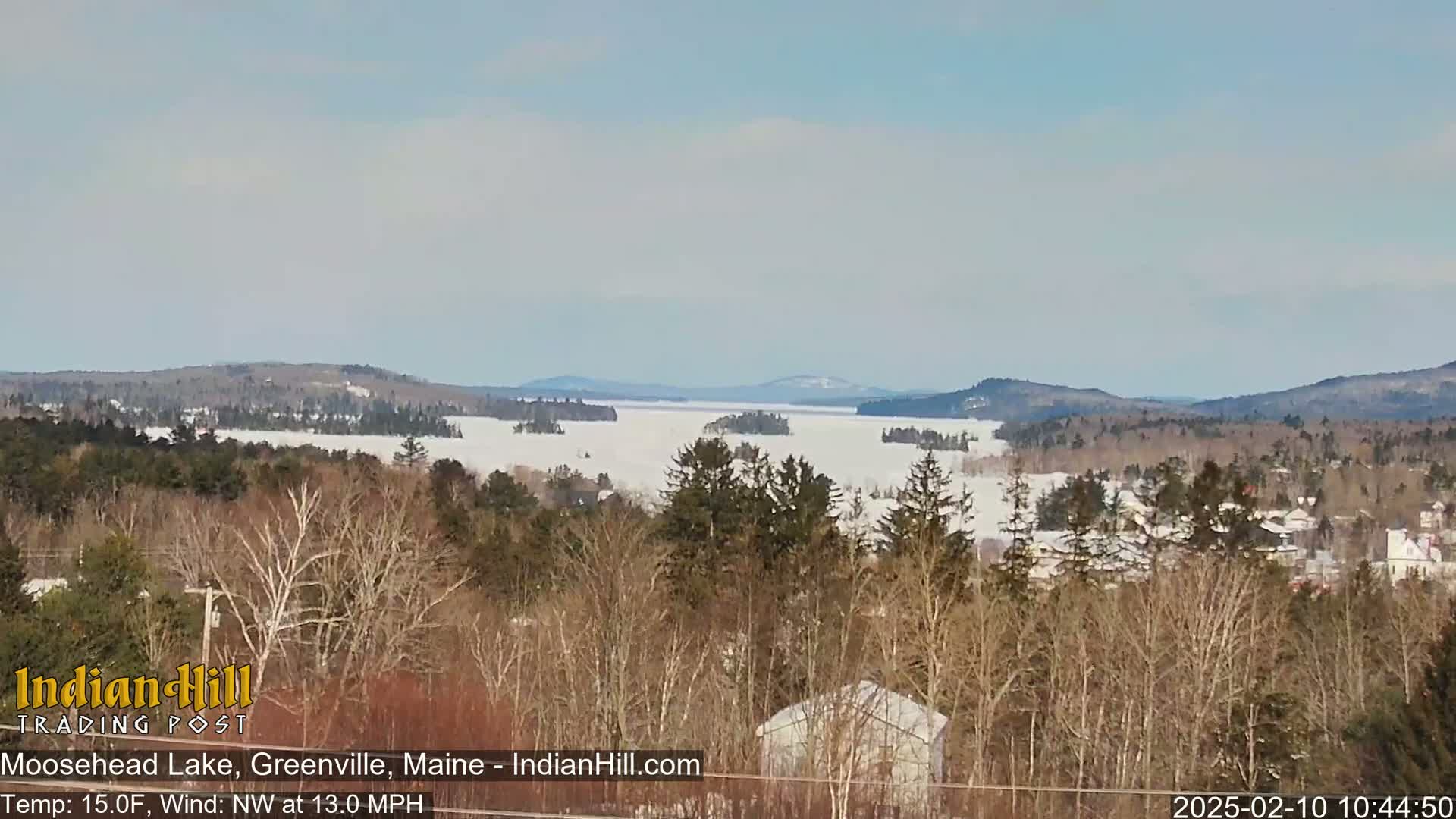 A mostly clear, cold, and windy day reveals a snow-covered Moosehead Lake, surrounded by bare winter trees and distant mountains.