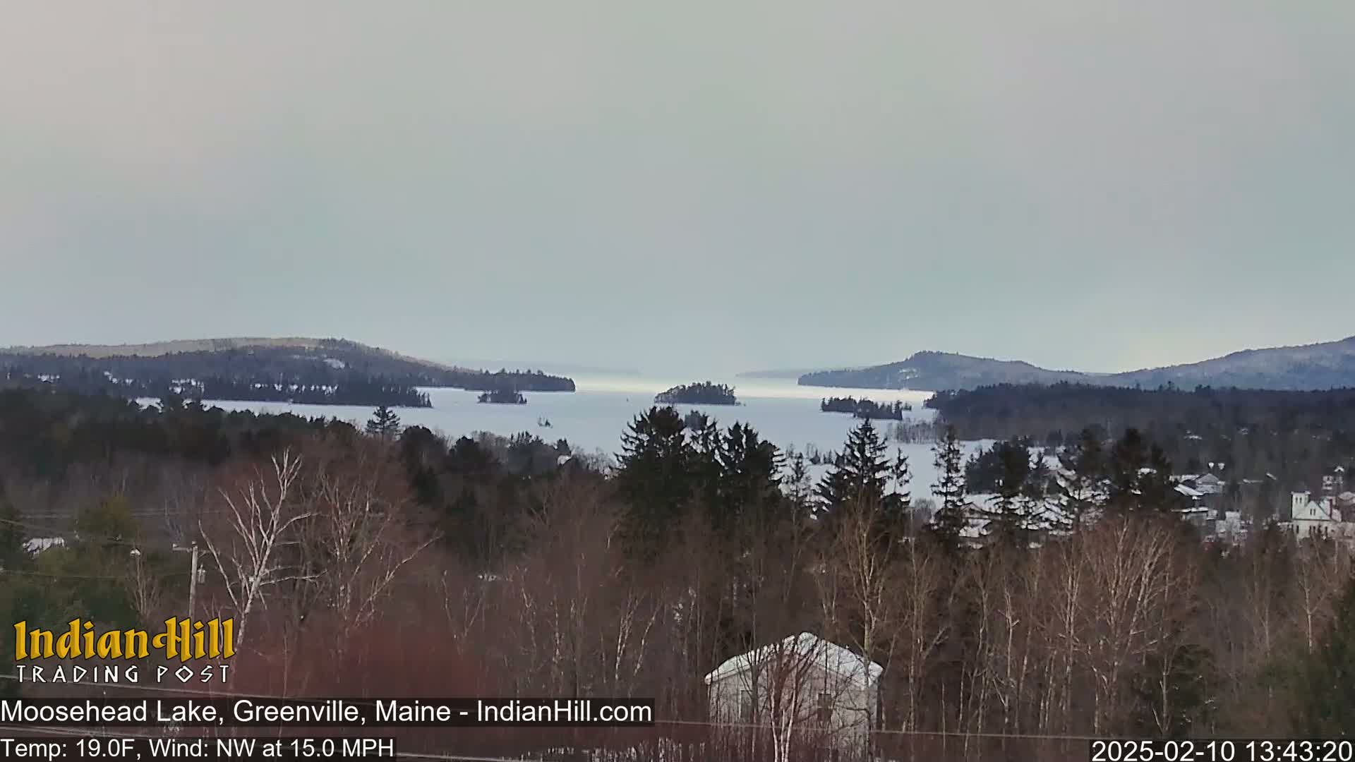 A snow-covered lake with several small islands is visible from a wooded hillside under an overcast sky, with a northwest wind blowing at 15 mph and a temperature of 19 degrees Fahrenheit.