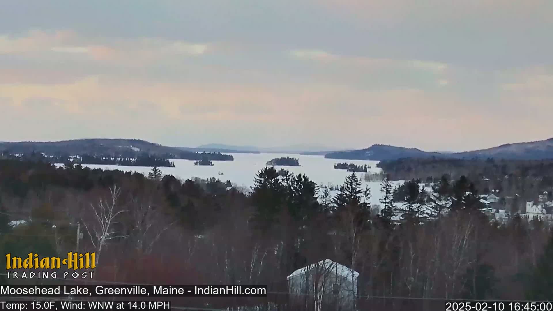 A snow-covered lake with several islands is seen from a high vantage point under a pale, overcast sky.
