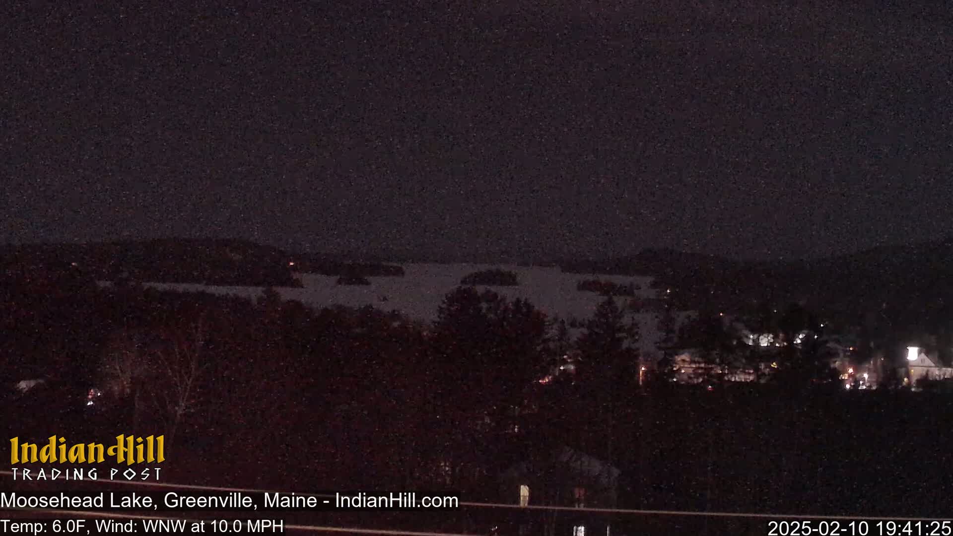 A dark, snowy night scene shows a body of water with islands in the distance, and a small town with faint lights nestled among dark trees in the foreground.