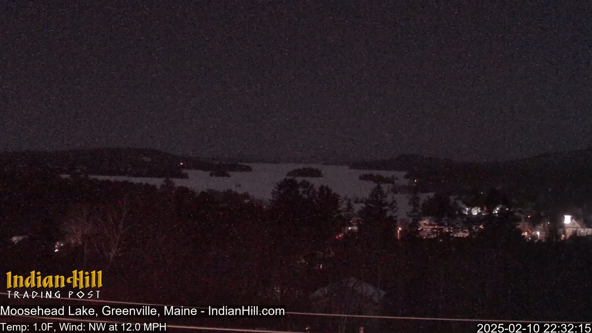A nighttime view of a snow-covered lake with sparsely lit buildings and trees in the foreground under a dark, possibly snowy sky.