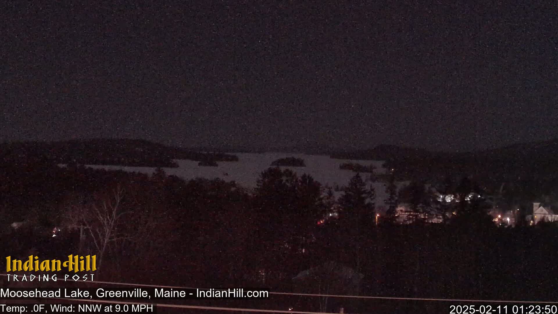A nighttime view of a snow-covered lake and a town under a lightly snowing sky.