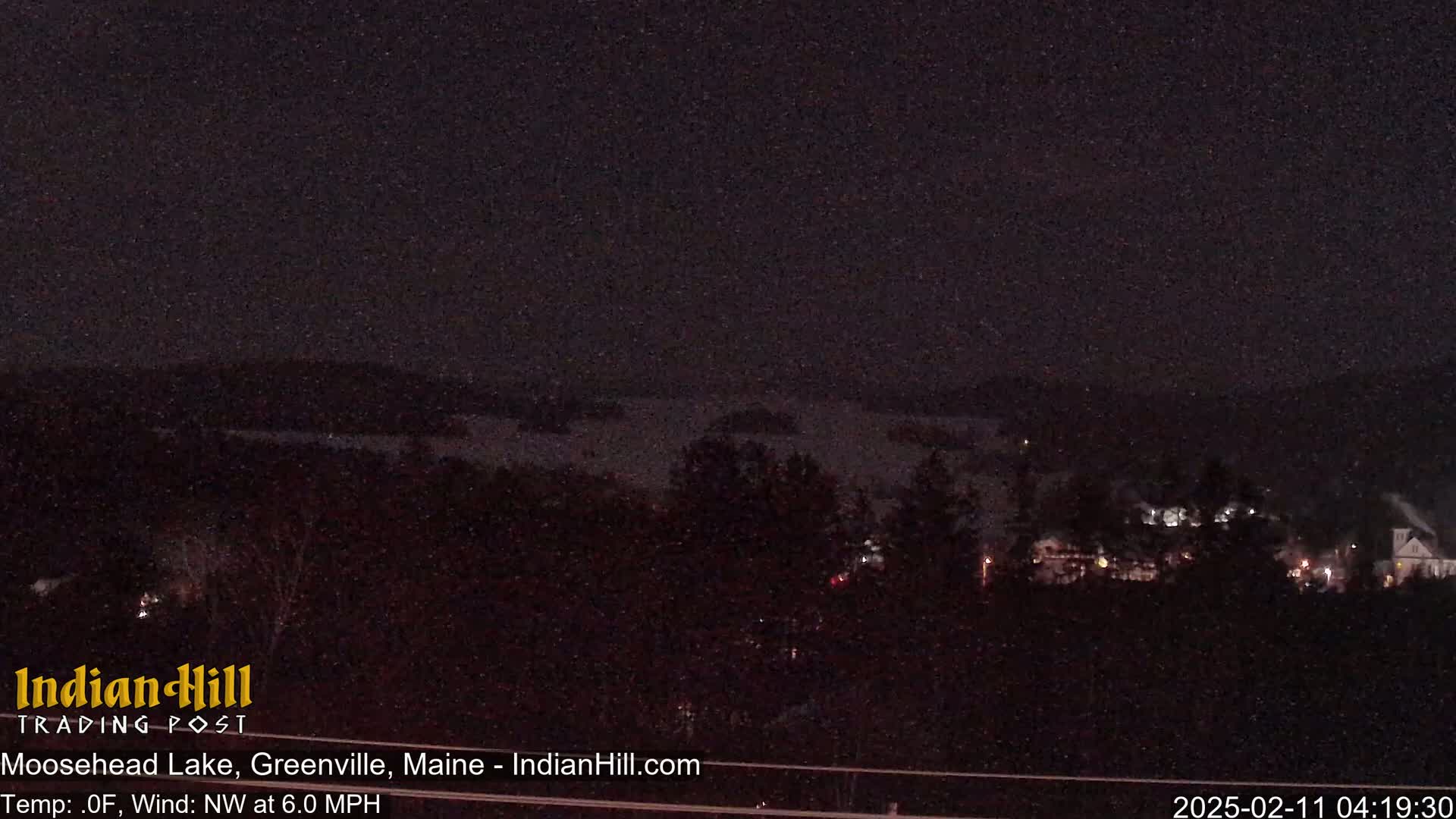 A nighttime view of a snow-covered lake and dark trees under a falling snow sky.