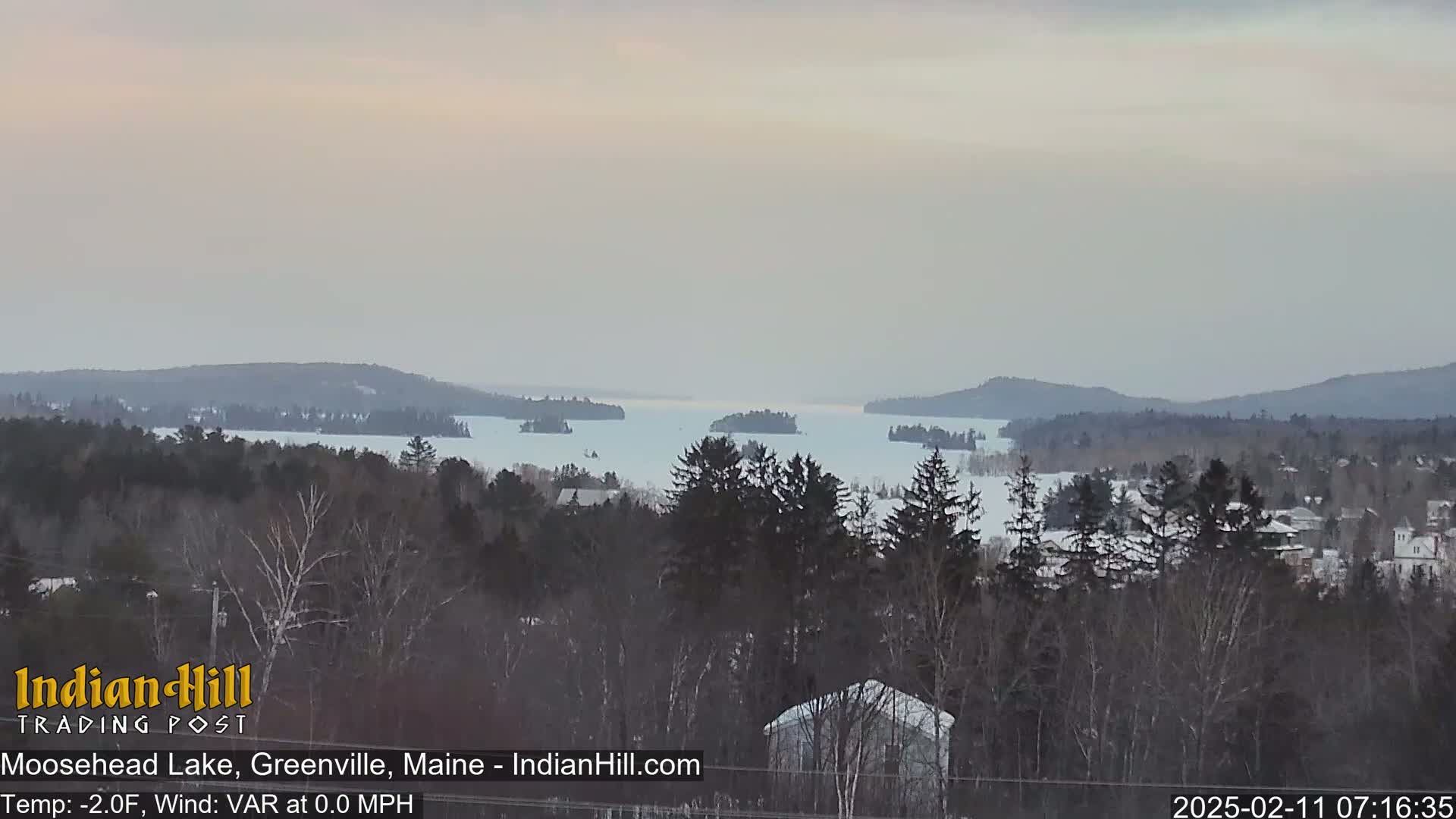 A snow-covered, partially frozen lake is visible from a wooded hillside, under an overcast sky.