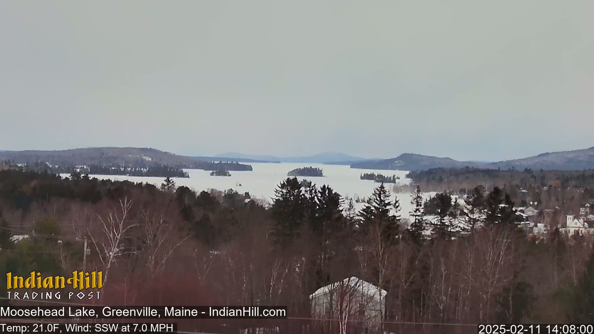 A mostly frozen lake stretches out under an overcast sky, surrounded by snow-covered trees and distant hills.