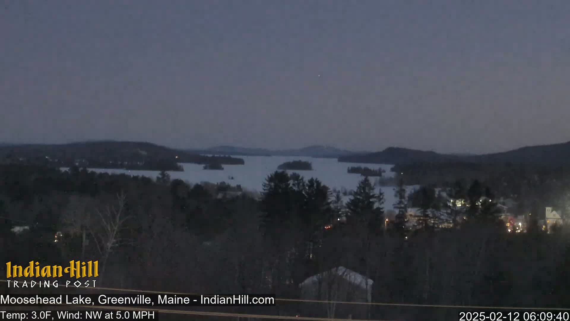 A twilight view of a snow-covered lake surrounded by dark, snow-dusted trees and hills under a dusky sky with a northwest wind.