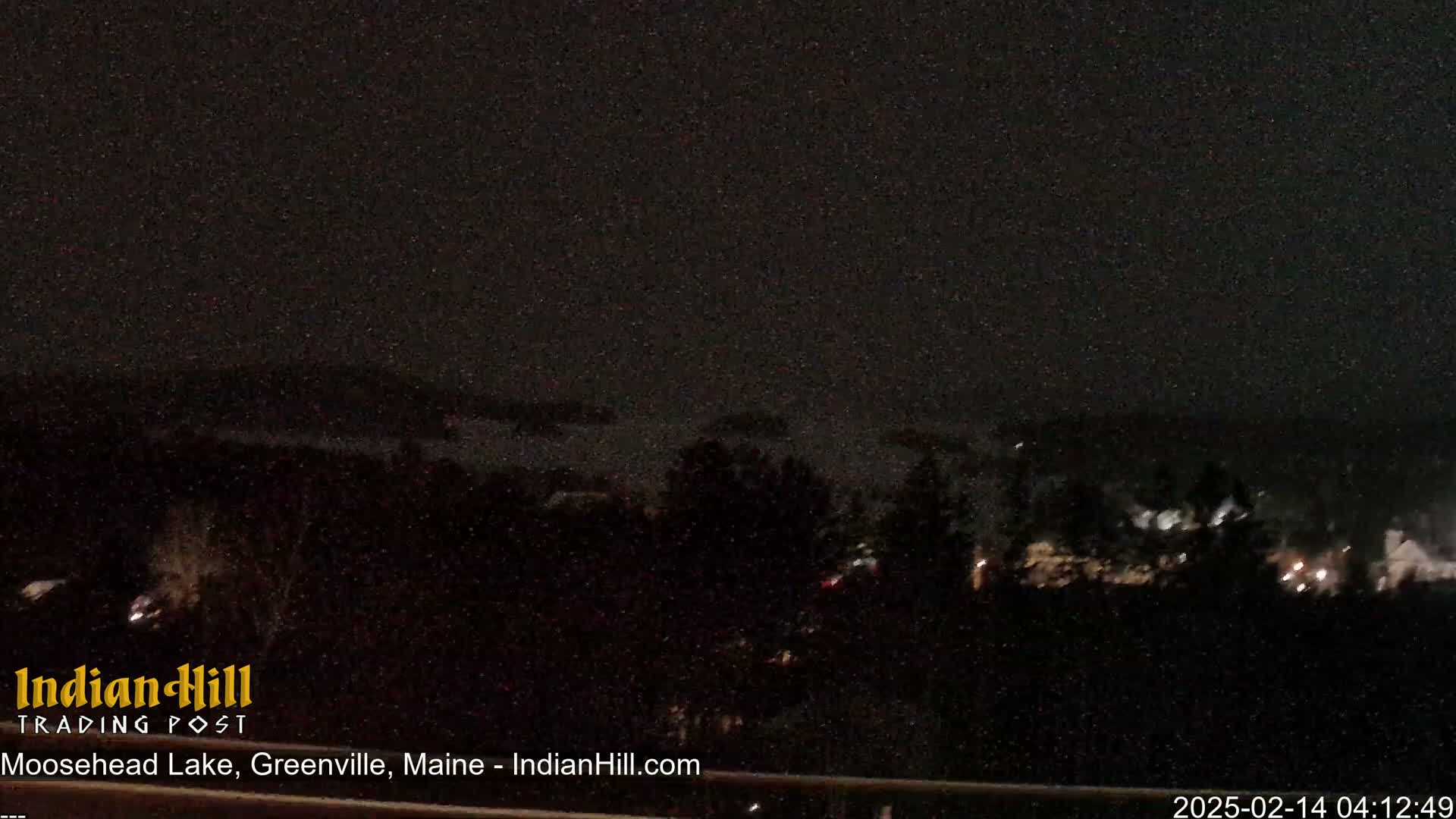 A nighttime, snowy view of a dark, wooded area with a few distant lights visible.