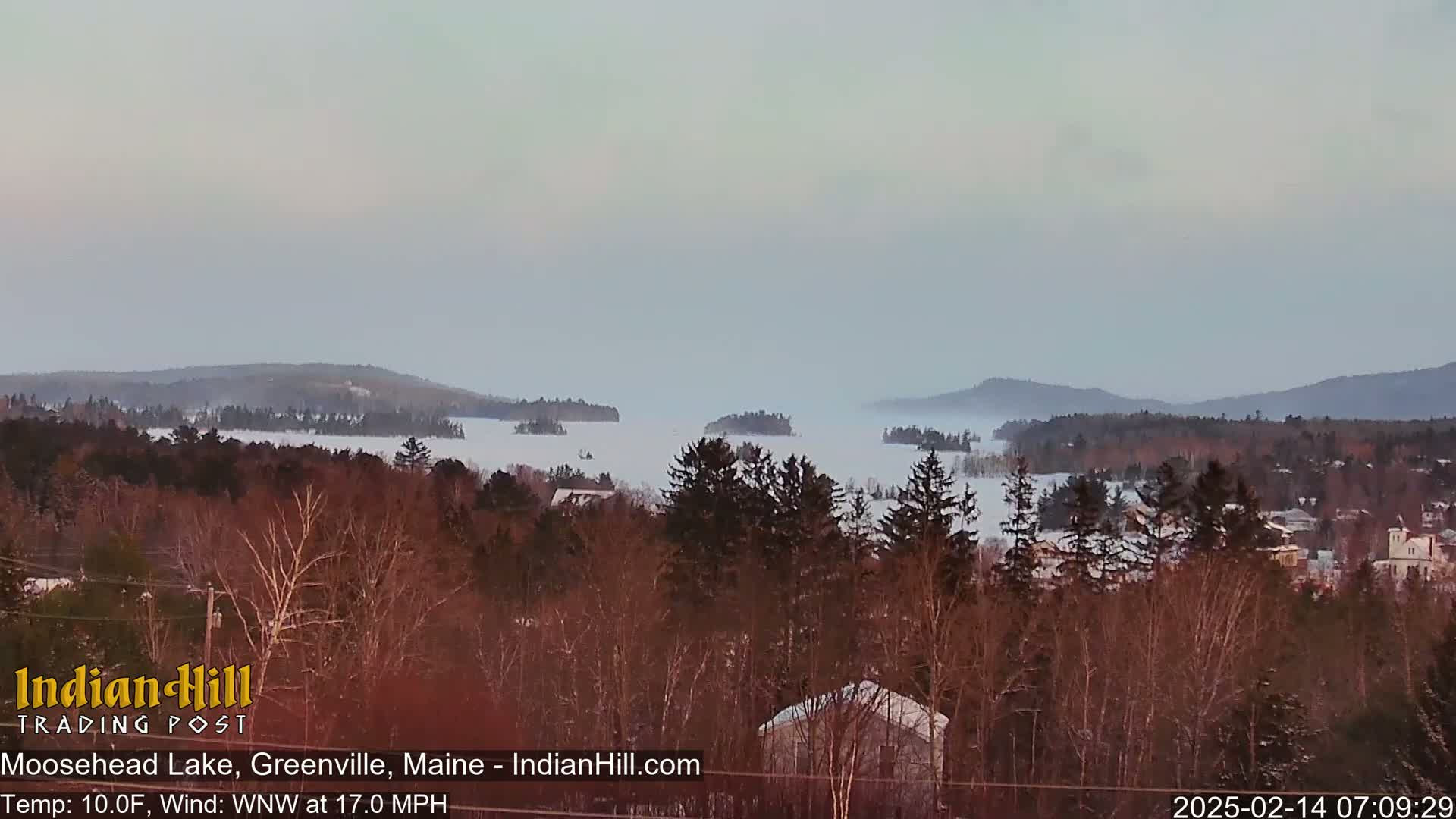 A snow-covered lake with numerous small islands is visible from a high vantage point, with a town and snow-covered forest in the foreground, under a hazy sky.