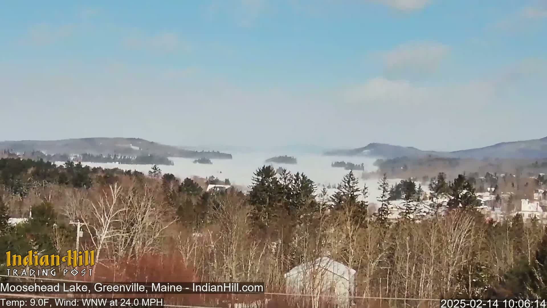A cold, windy day shows a lake shrouded in fog, with snow-dusted trees and distant hills visible in the background.