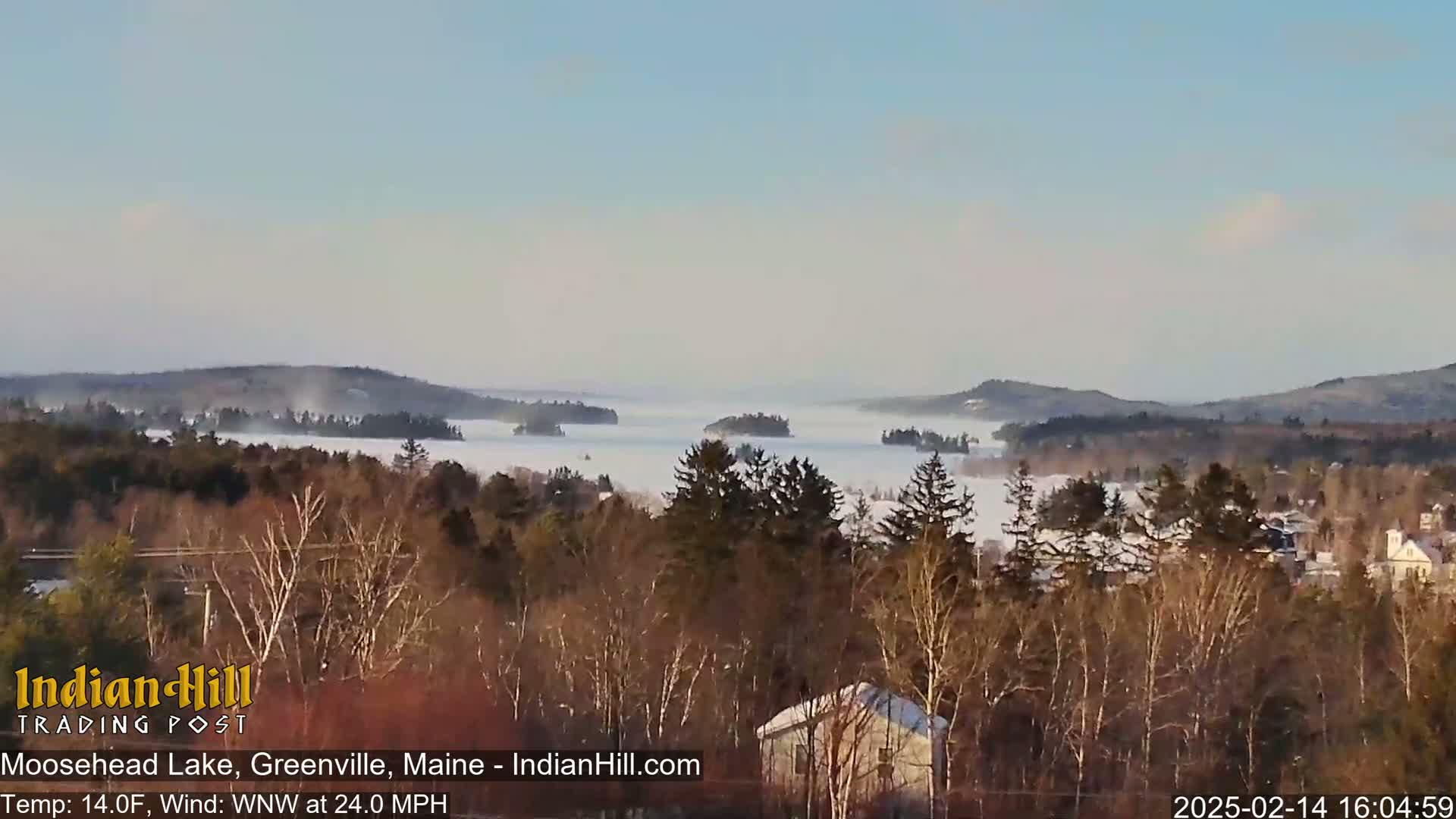 A snow-covered lake is partially obscured by fog under a clear sky, viewed from a wooded hillside with a light dusting of snow on the trees and houses in the foreground.