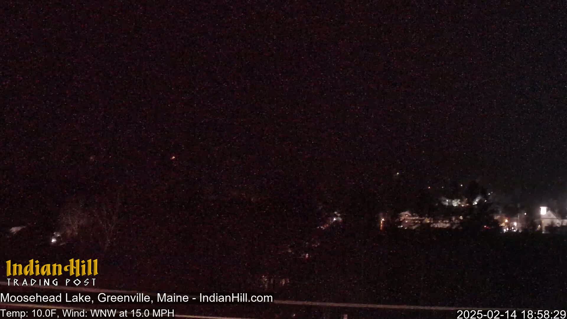 A nighttime, low-light view of a dark landscape with a few distant lights, under a clear, starry sky with a 10°F temperature and 15 mph wind from the west-northwest.
