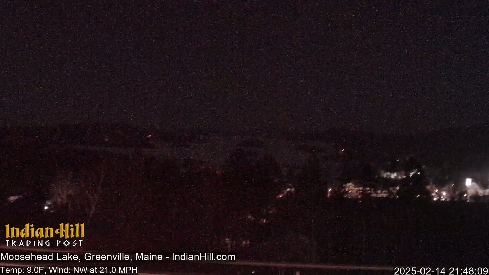 A nighttime view of a dark lake and shoreline with faint lights visible in the distance, under a dark sky with a northwest wind at 21 mph and a temperature of 9 degrees Fahrenheit.