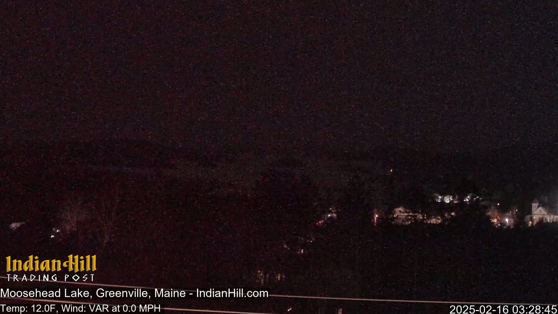 A nighttime view of a dark, snow-covered landscape with a few faint lights visible in the distance under a mostly clear sky.
