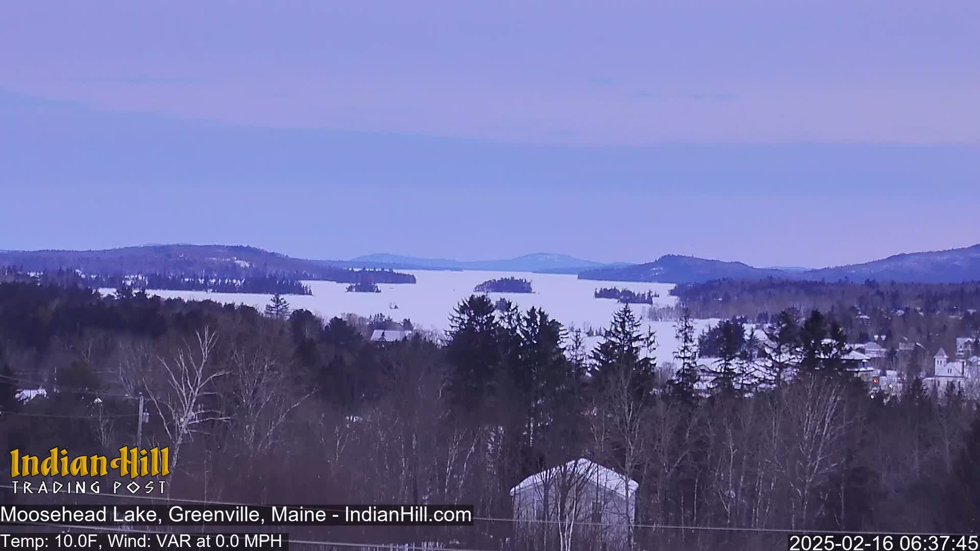 A snow-covered lake dotted with islands is seen from a snowy, wooded hillside under a pale purple sky.