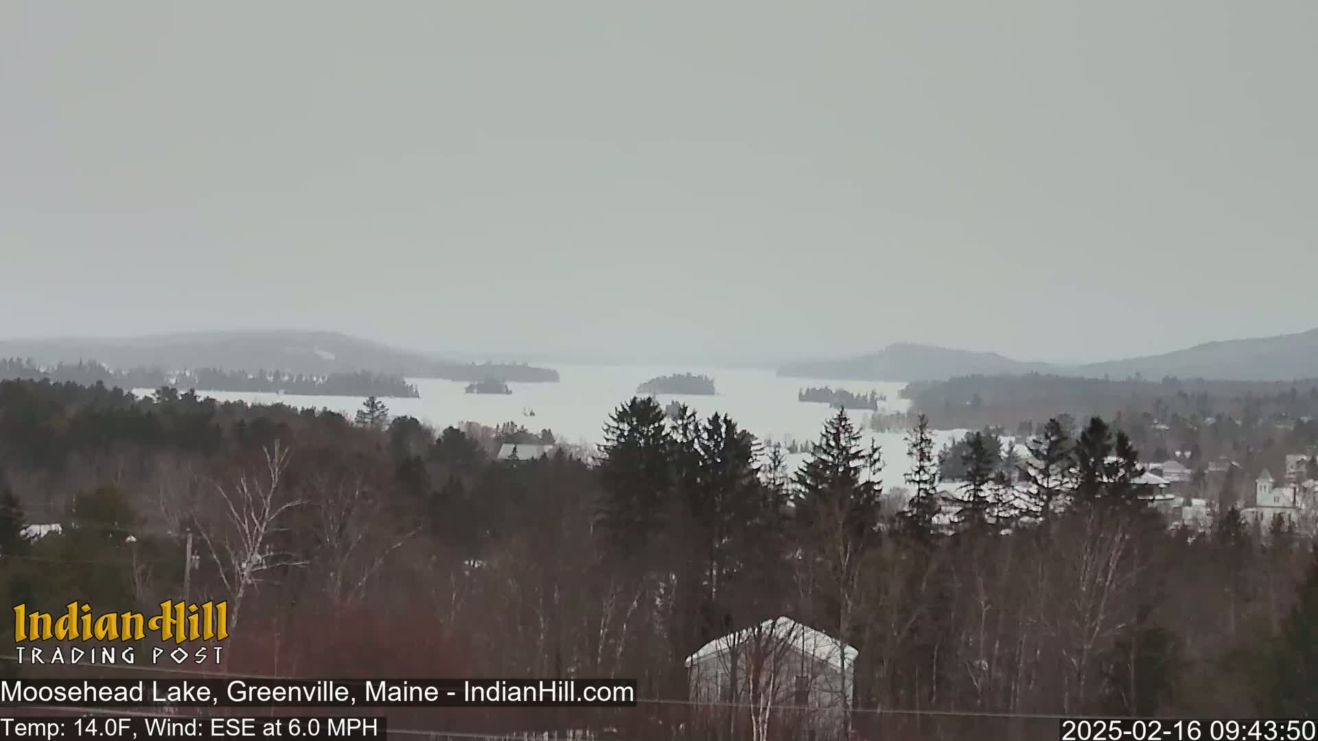 A snow-covered lake stretches to distant, snow-dusted mountains under an overcast sky.